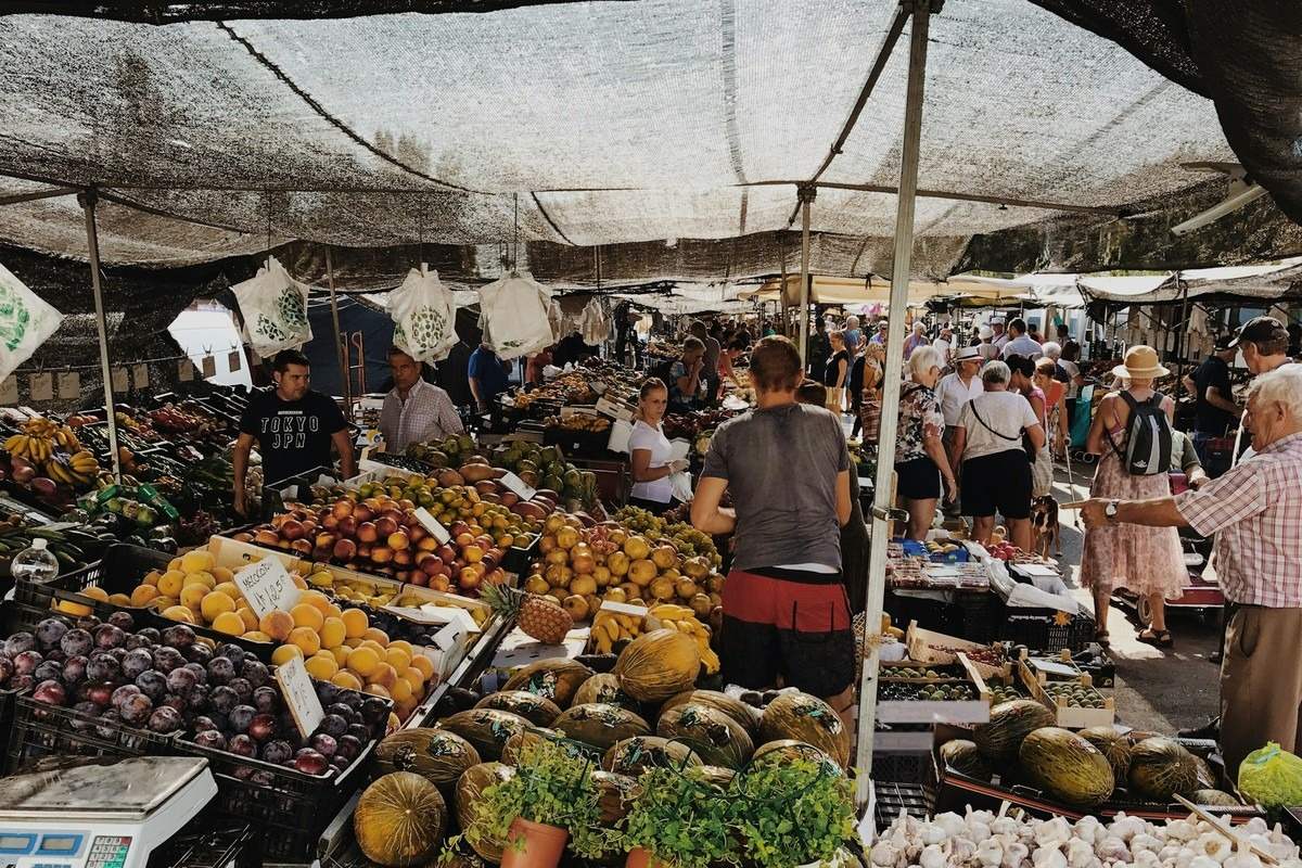Gölgelik altında yan yana dizilmiş tezgâhlarda farklı satıcıların ürünlerini sattığı kalabalık bir açık pazar